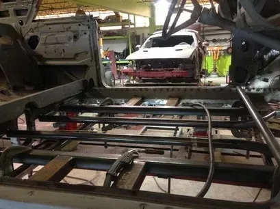 Car frame assembly inside a workshop, with a white car body in the background and metal supports in the foreground