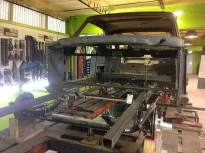 Disassembled car chassis in a workshop, with exposed frame and engine bay under bright green lights.