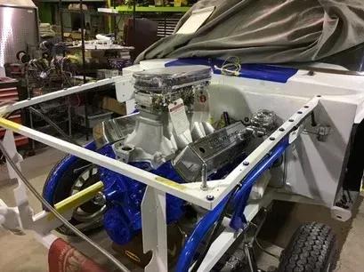 Race car chassis with exposed engine and blue accents in a workshop, partly covered by a tarp