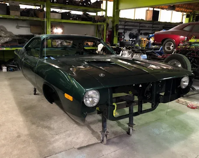 Dark green classic car in a workshop, missing front bumper and wheels, with a red car in the background
