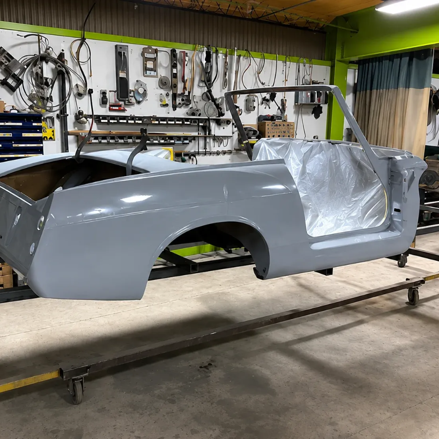 Gray classic car body shell on a workbench in an auto restoration workshop, with tools on the wall.