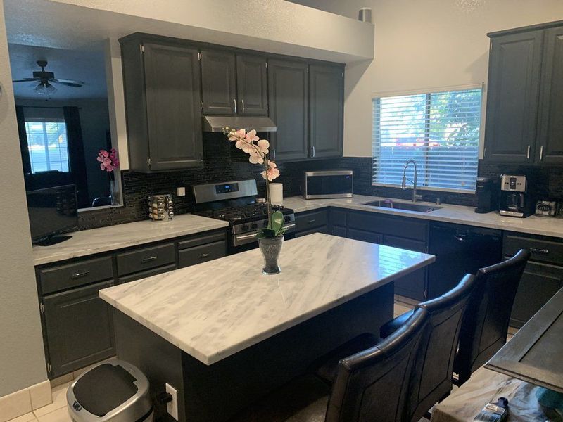 Modern kitchen with dark cabinets, white island, and black chairs by a window.