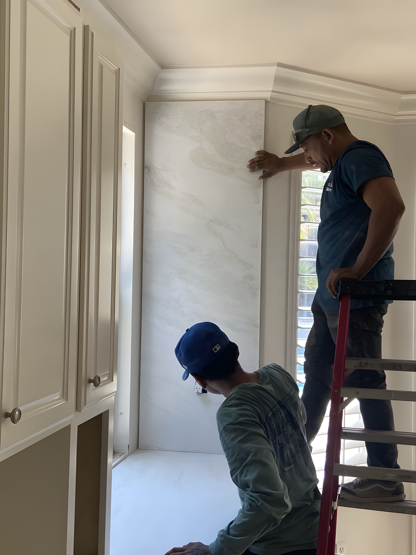Two workers install wallpaper in a bright kitchen, one on a ladder and one below.