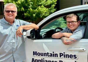 Two men leaning on a white Mountain Pines Appliances truck, smiling outdoors.