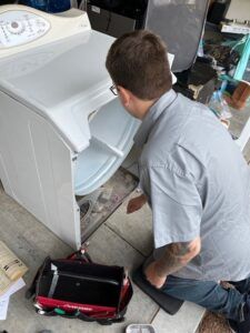Technician kneeling beside a white appliance, inspecting its open front panel with tools nearby.