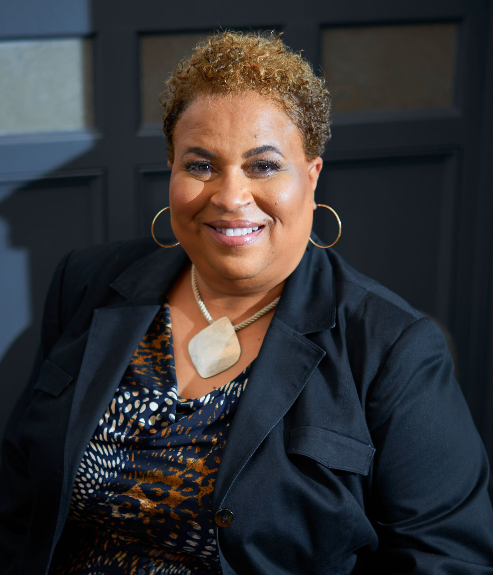 A professional portrait of a person with short curly hair, wearing a blazer, patterned top, and a large stone necklace.