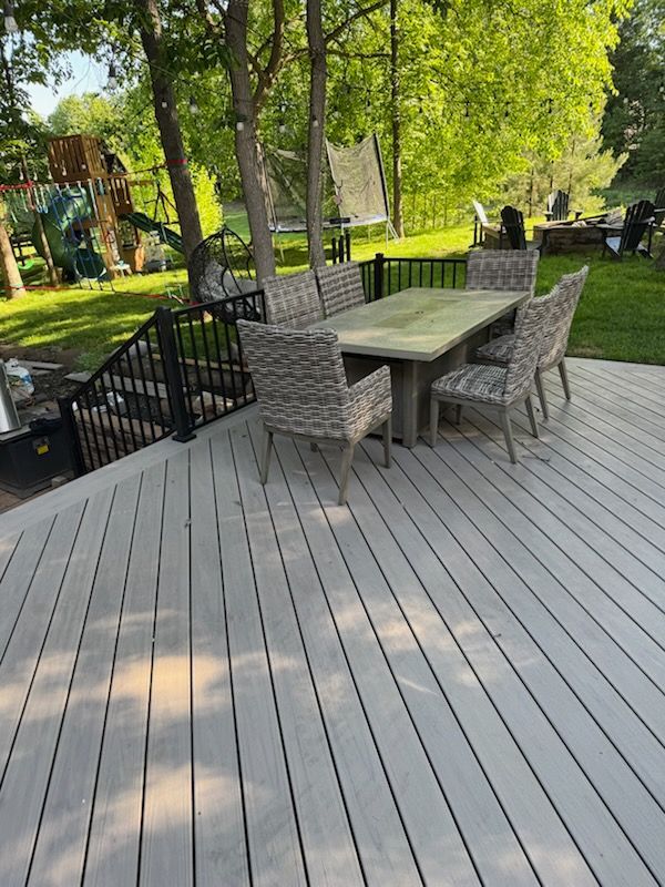 Outdoor deck with a table and wicker chairs, overlooking a green backyard with trees and a black fence.