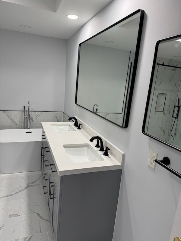 Modern bathroom with double vanity, large mirrors, gray cabinets, and a white bathtub with marble walls
