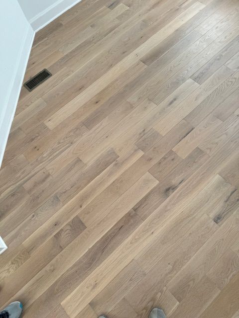 Light wood flooring in an empty room with a floor vent and white wall trim