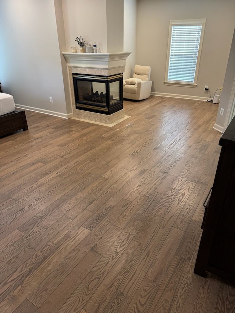 Empty living room with hardwood floors, corner fireplace, white chairs, and a window with blinds
