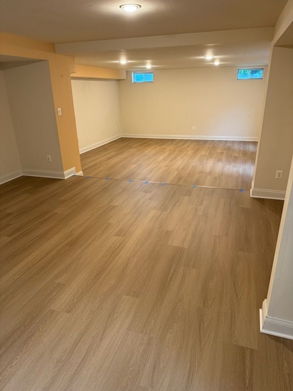 Empty finished basement with wood-look flooring, beige walls, and small high windows with blue light.