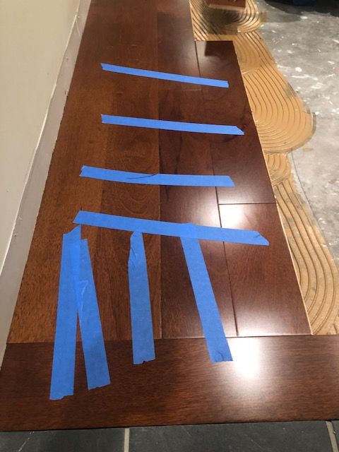 Blue tape strips arranged on a wooden floor near a rug and wall.
