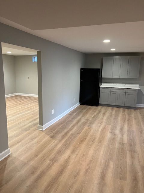 Empty modern apartment living area with light wood floors, gray walls, and a small kitchen with black fridge.