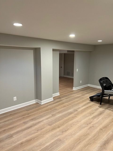 Empty gray room with wood-look flooring, recessed lights, and a black chair near a doorway.