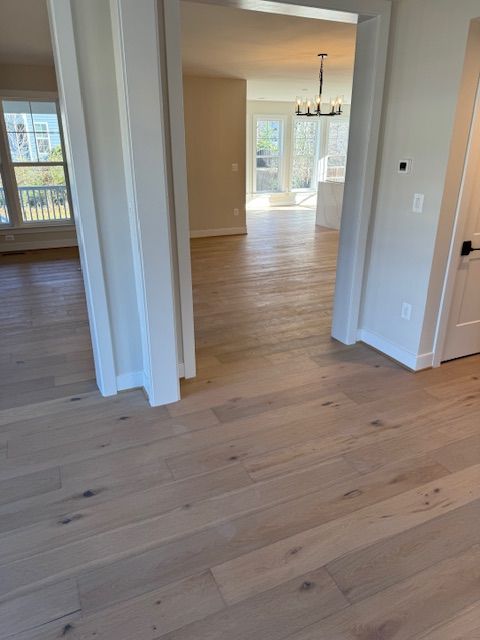 Empty modern room with light wood floors, white trim, and a dining area visible through an open doorway.
