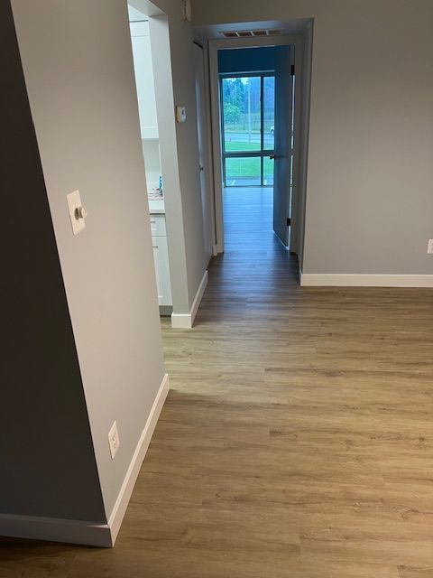 Empty hallway with gray walls, light wood floors, and a view of a glass door at the end.