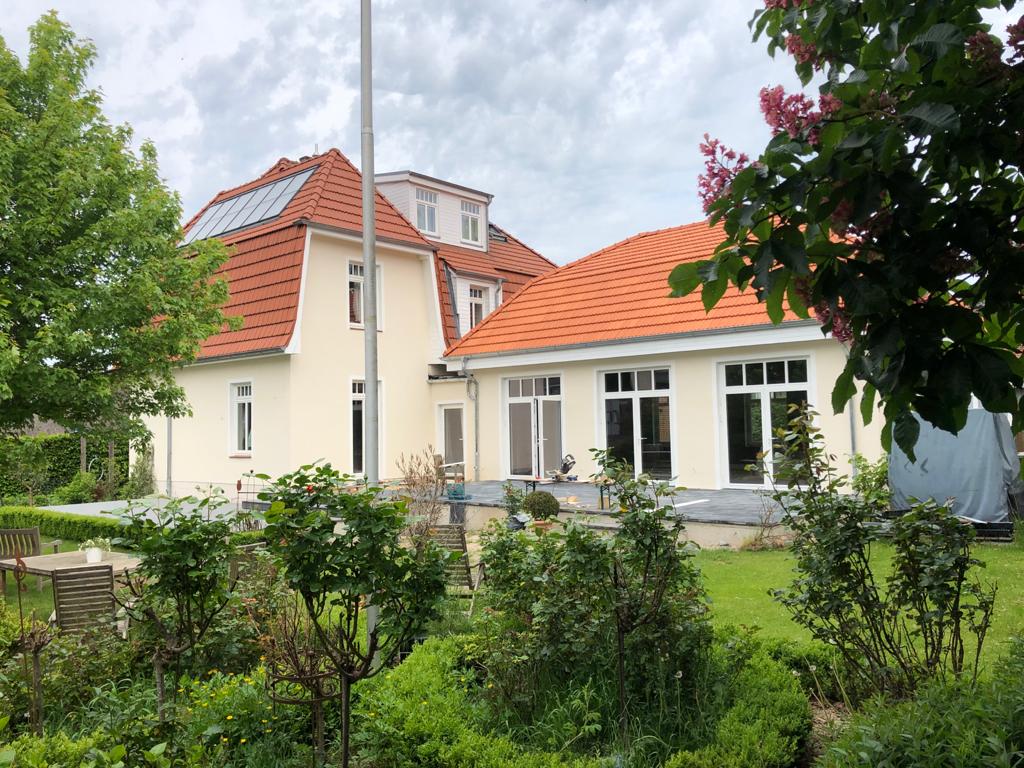 Ein großes weißes Haus mit rotem Dach ist von Bäumen und Büschen umgeben.