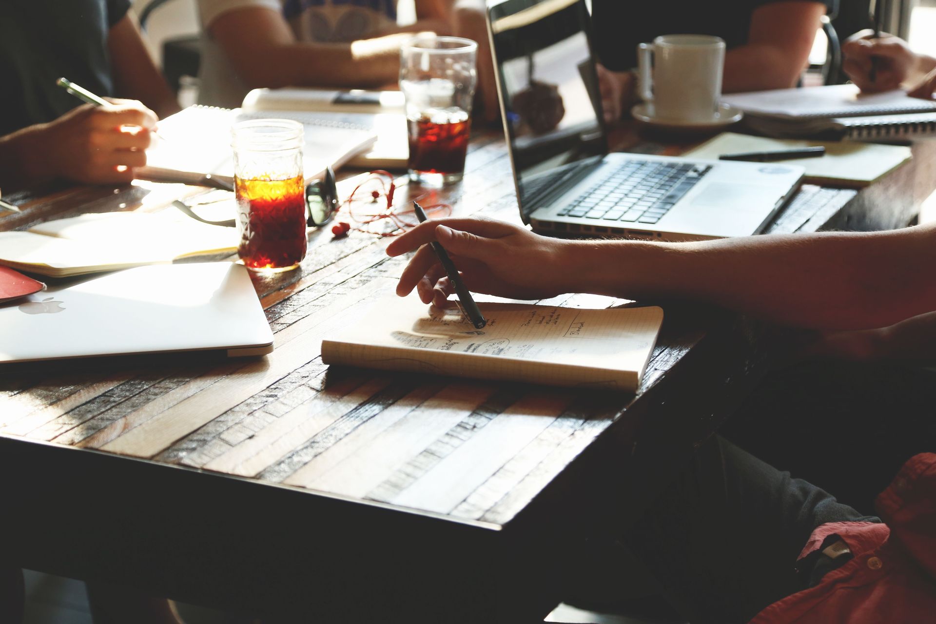 People working at a sunlit table with notebooks, laptops, and drinks during a writing workshop