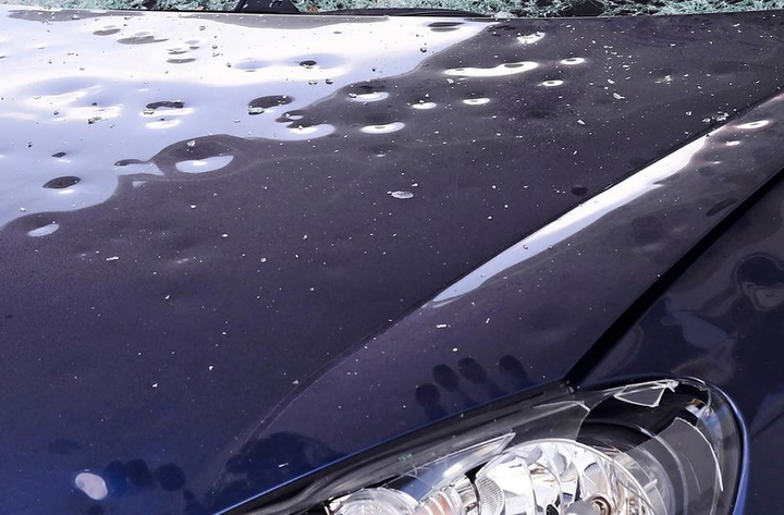 Cofano di un'auto blu scuro con gocce d'acqua e un faro visibile in basso a destra.