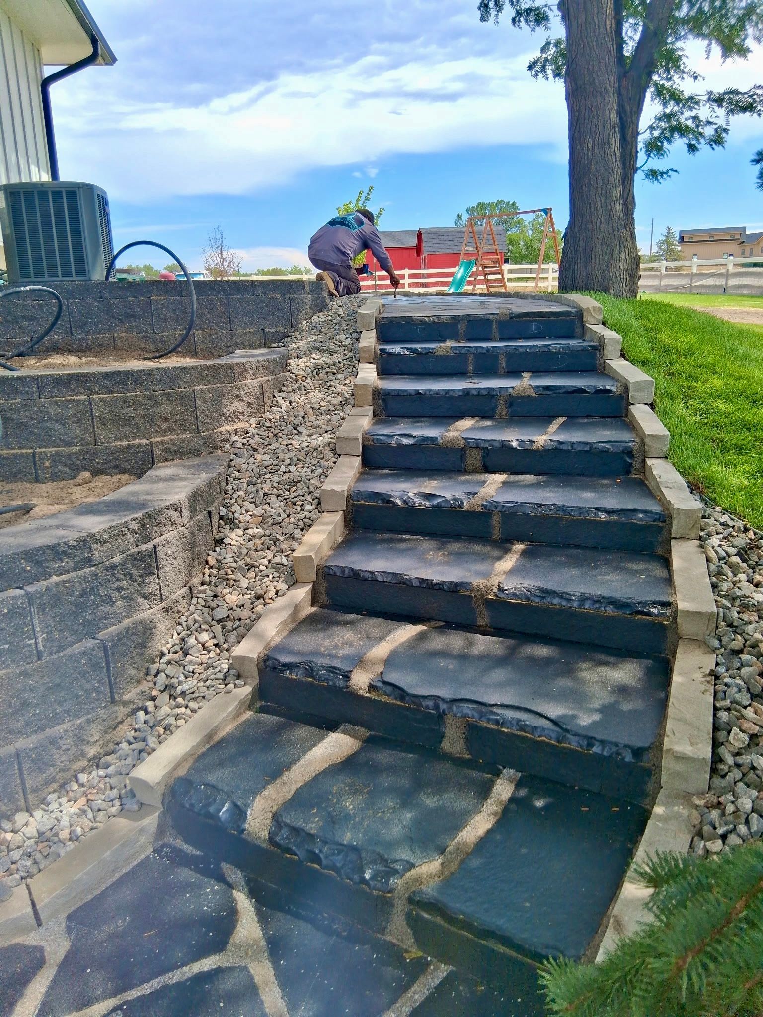 Stone steps in a park beside a stone wall, with people sitting at the top near a playground and grassy slope.