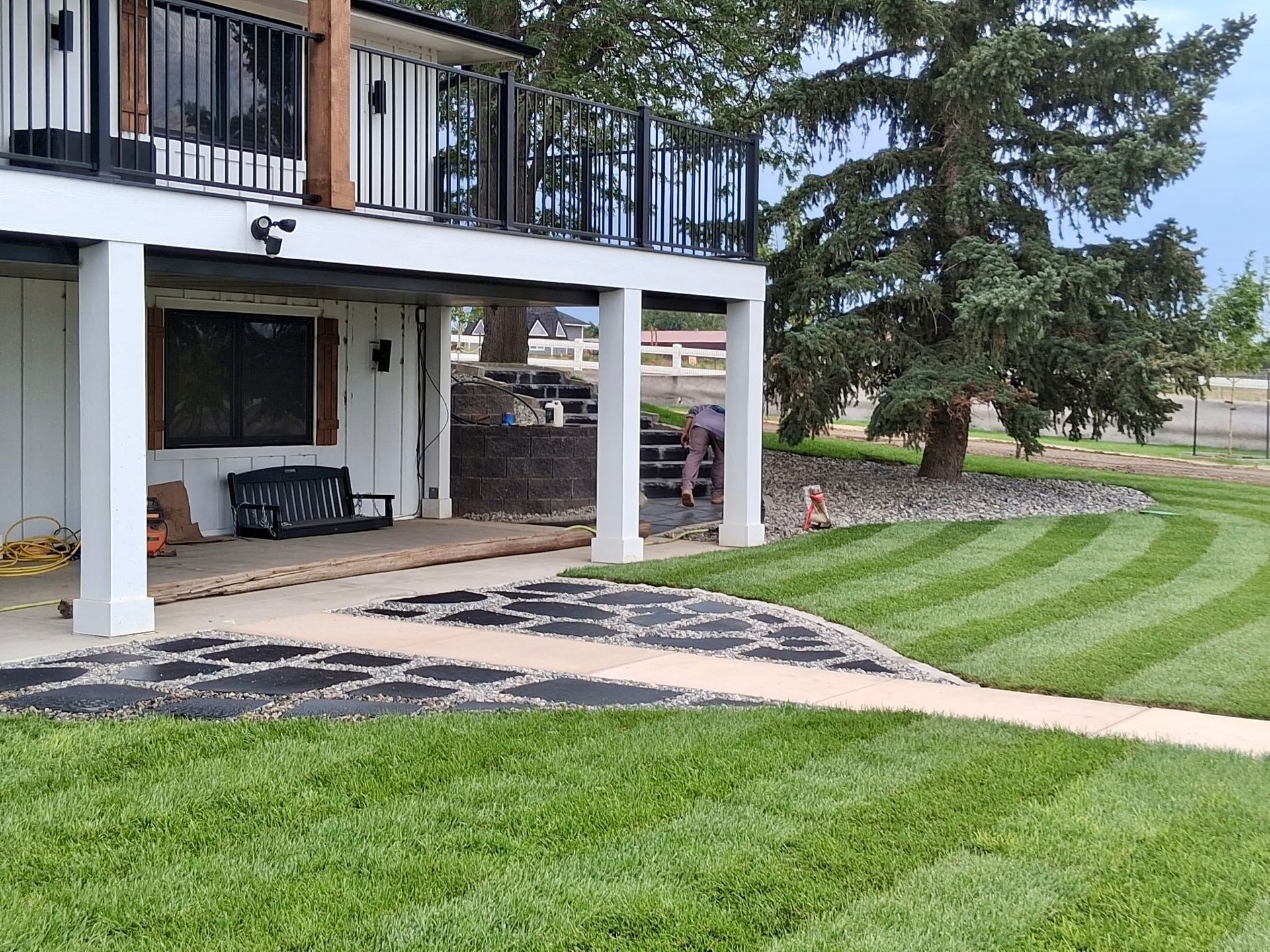 Backyard patio beneath a raised deck beside a striped green lawn and large trees