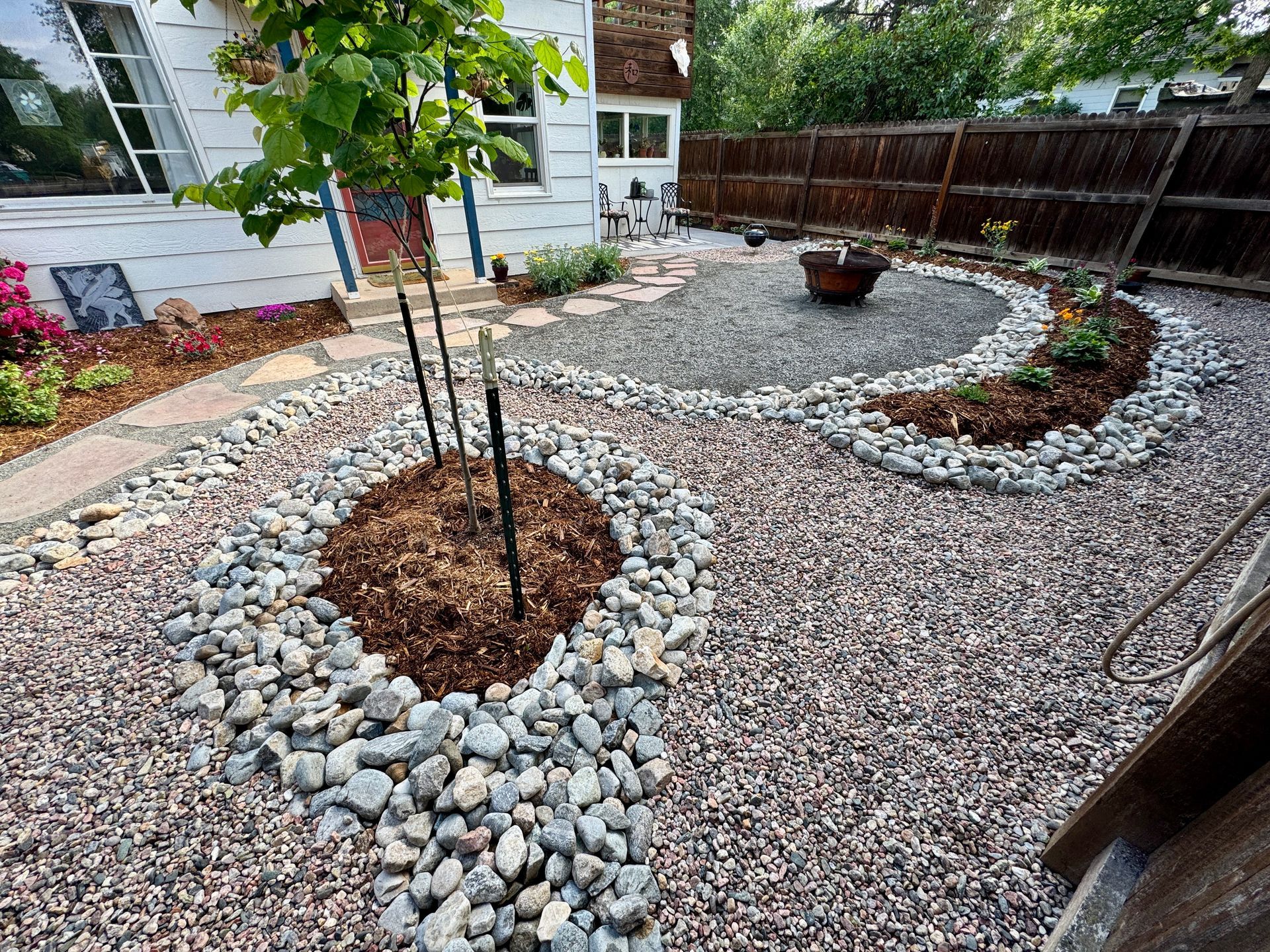 Gravel backyard with a young tree, landscaping stones, fire pit, and wooden fence.