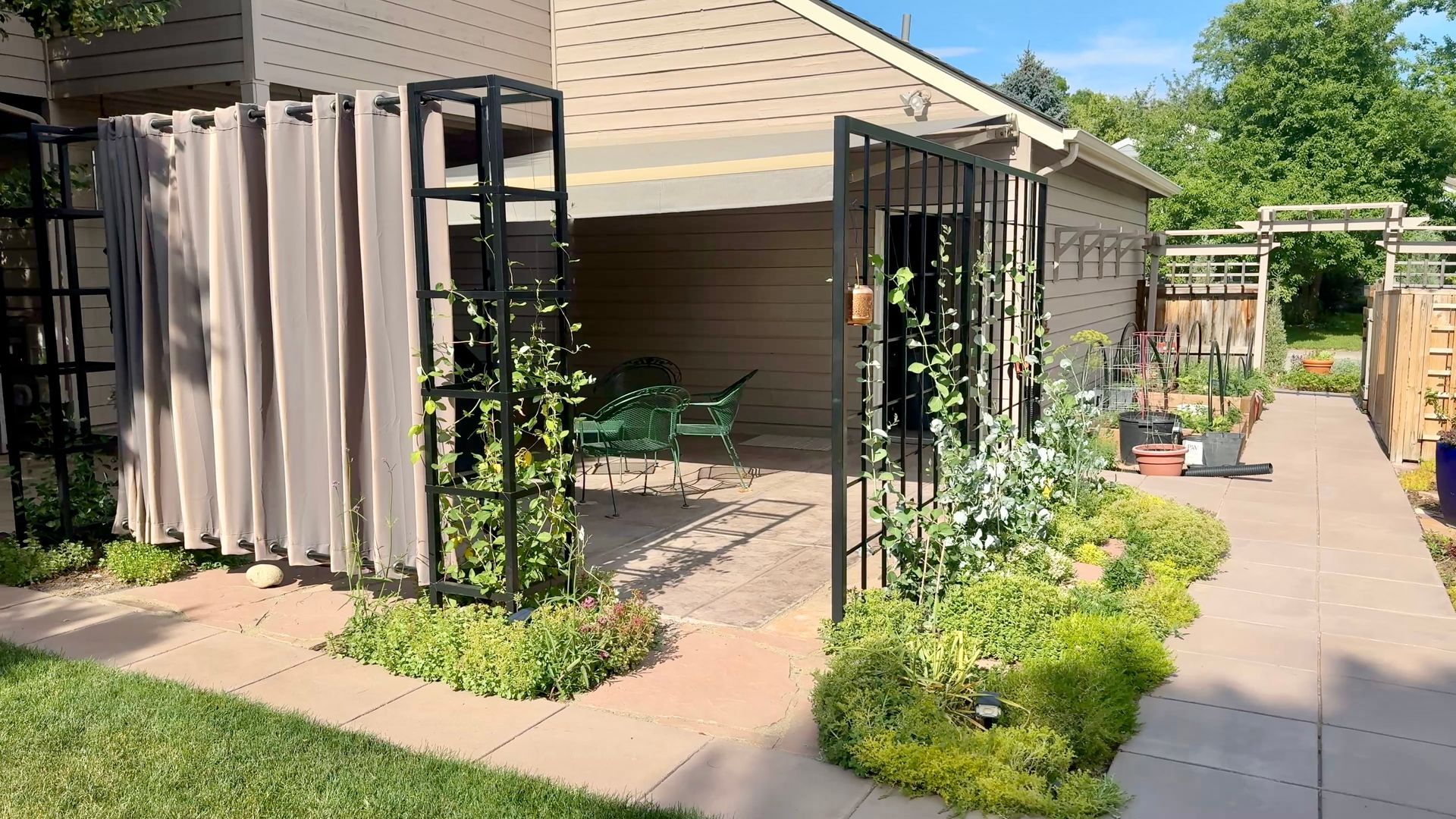 Garden patio with black metal trellises, potted plants, and a sunlit walkway beside a house