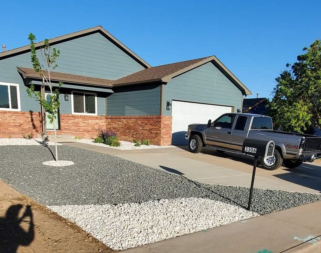 House with gray and red brick exterior, a driveway, and a pickup truck. Gravel landscape with small tree.