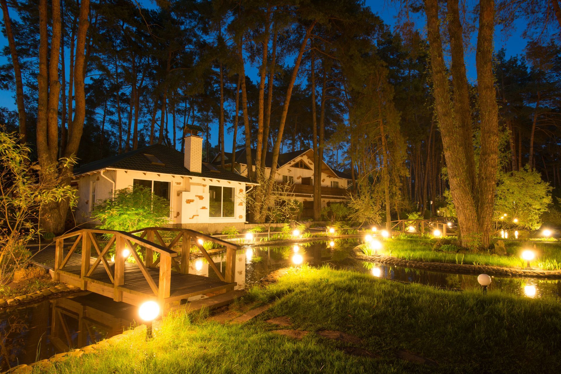 A night scene with a white building, lit footbridge, pond with lights, and tall trees.