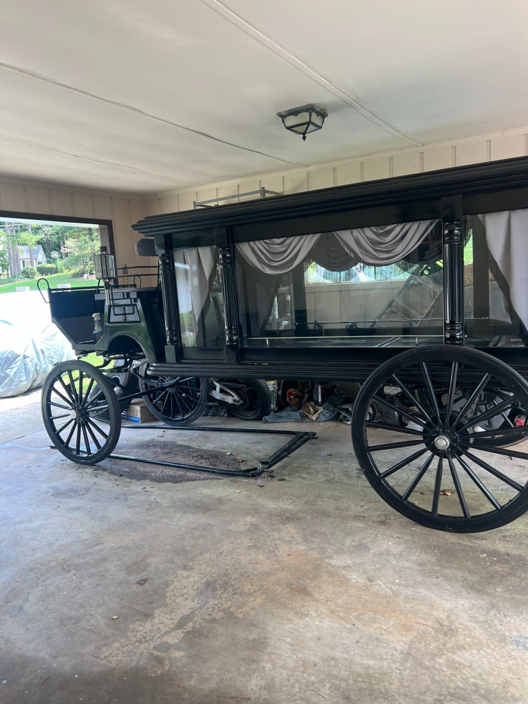 Vintage black horse-drawn hearse in a garage, with large spoked wheels and draped windows.