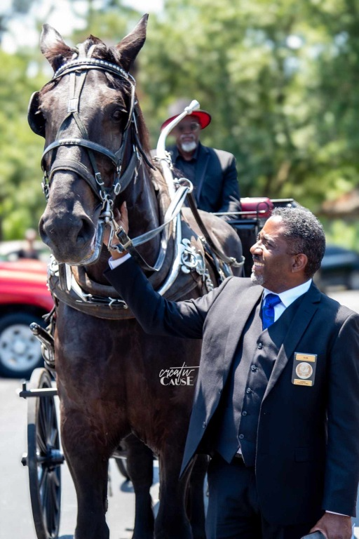 A mounted rider in a red hat beside a dark horse, with a man in a suit standing in the foreground.