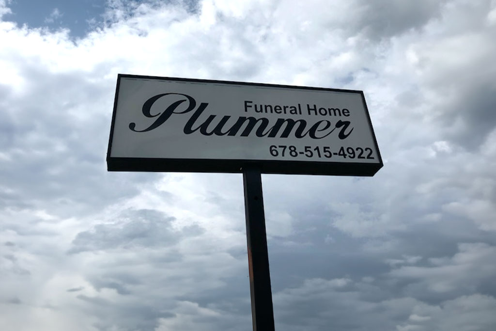 Plummer Funeral Home sign on a pole against a cloudy sky, with phone number 678-945-7922.