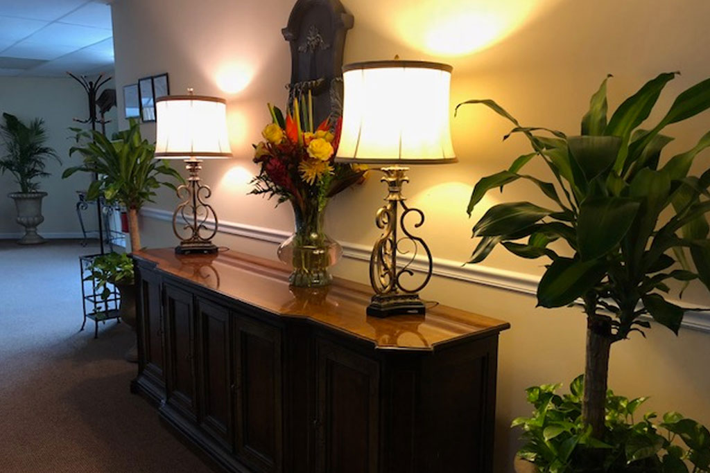 Dim hallway console table with lamps, flowers, and potted plants against a beige wall