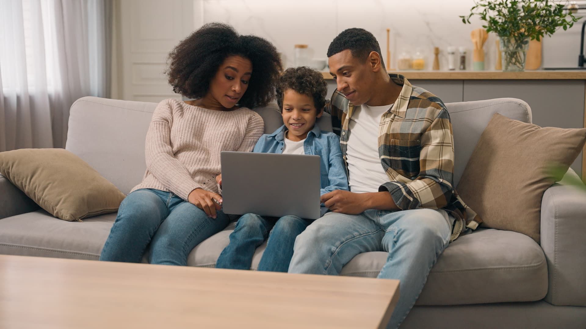 Family sitting on a couch, smiling while looking at a laptop in a bright living room.