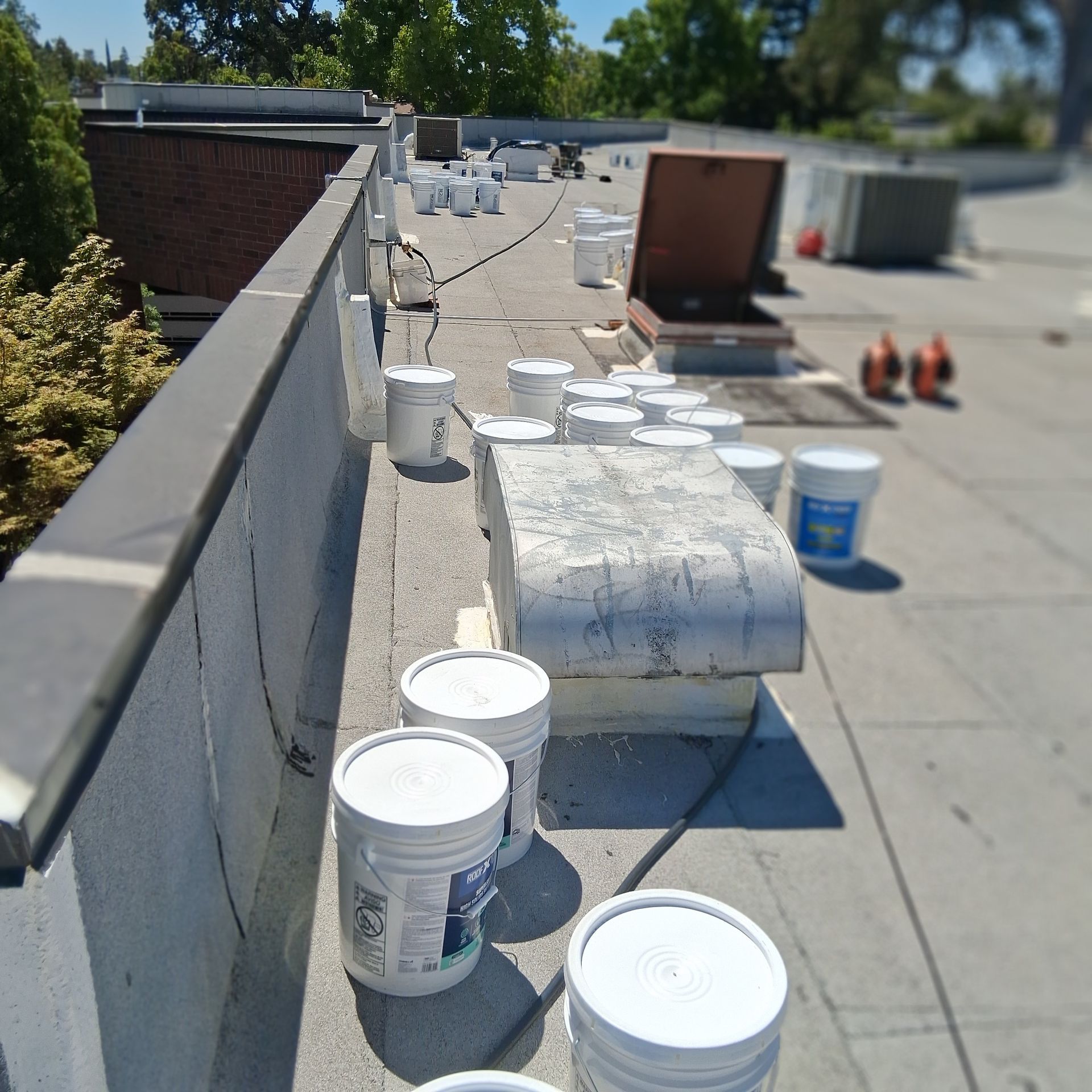 Rooftop with buckets, rolls of roofing material, and tools along a parapet wall under bright sun