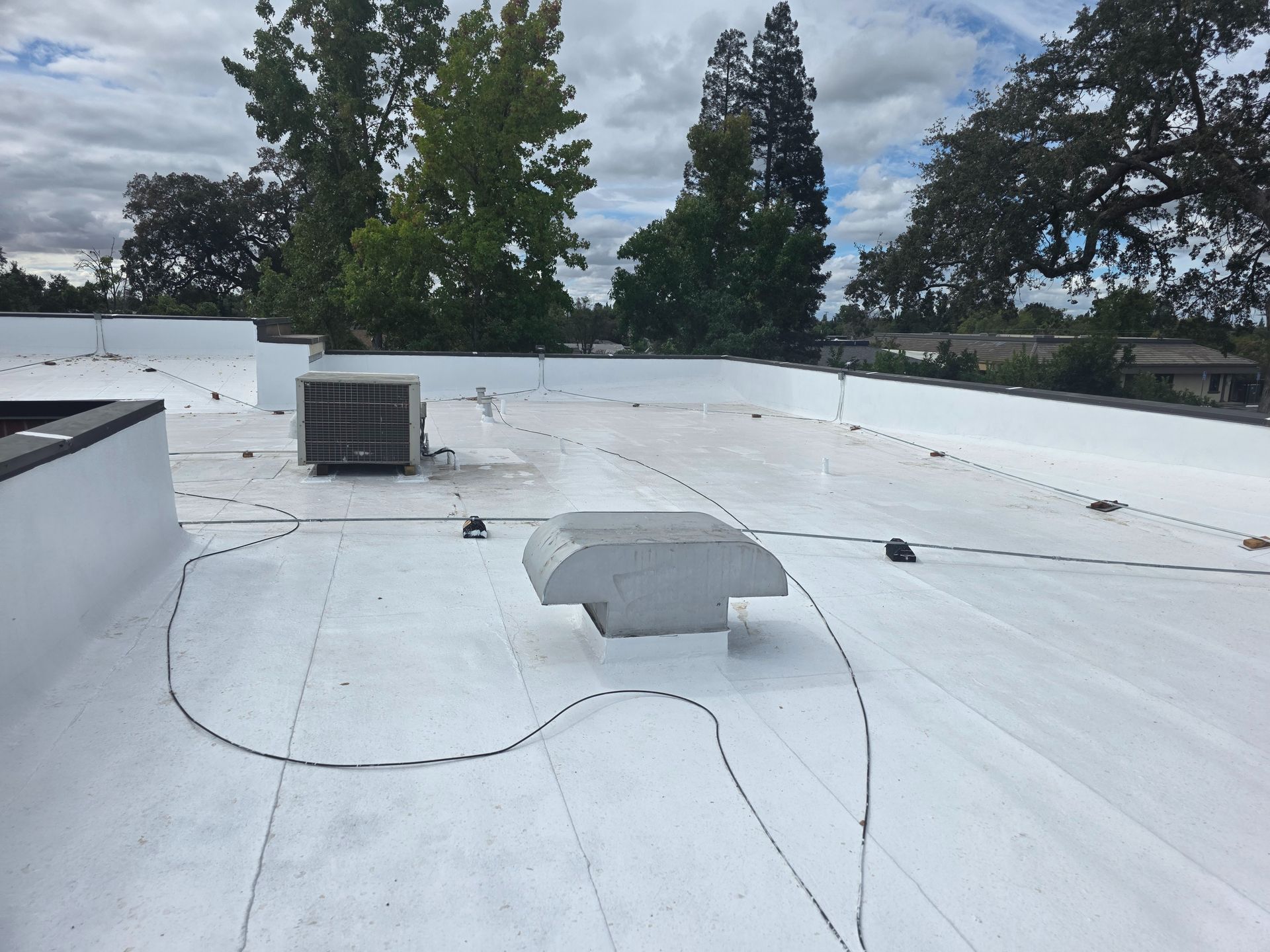 White flat rooftop with vents and cables under a cloudy sky, trees visible beyond the roofline