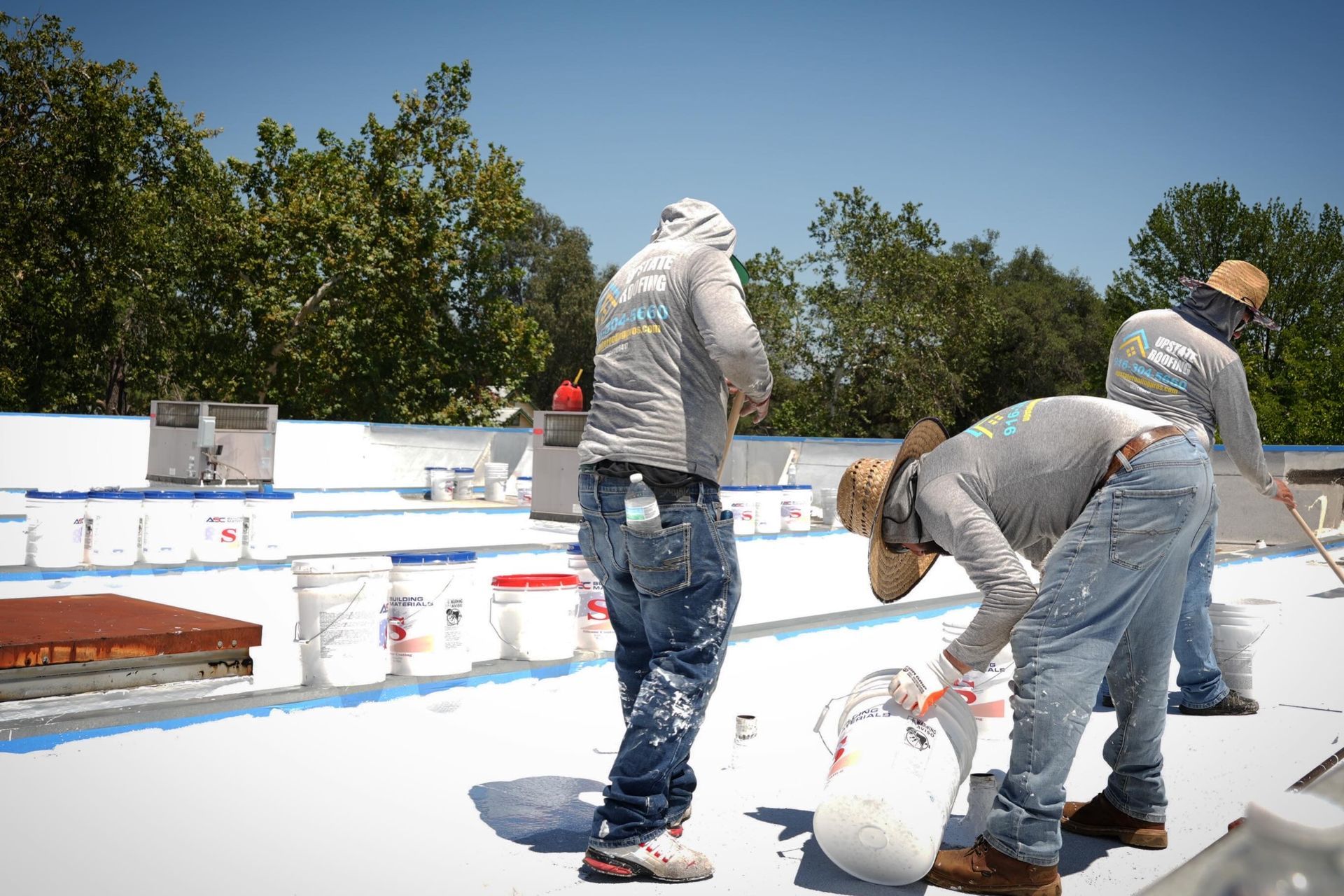Workers painting a white rooftop on a sunny day, with buckets and tools nearby.