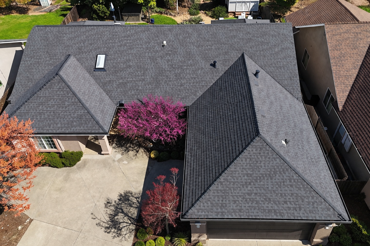 Aerial view of a house with dark gray roofs, a light driveway, and pink flowering shrubs in the yard.