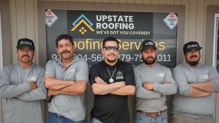 Five workers pose with arms crossed in front of an Upstate Roofing sign and storefront.