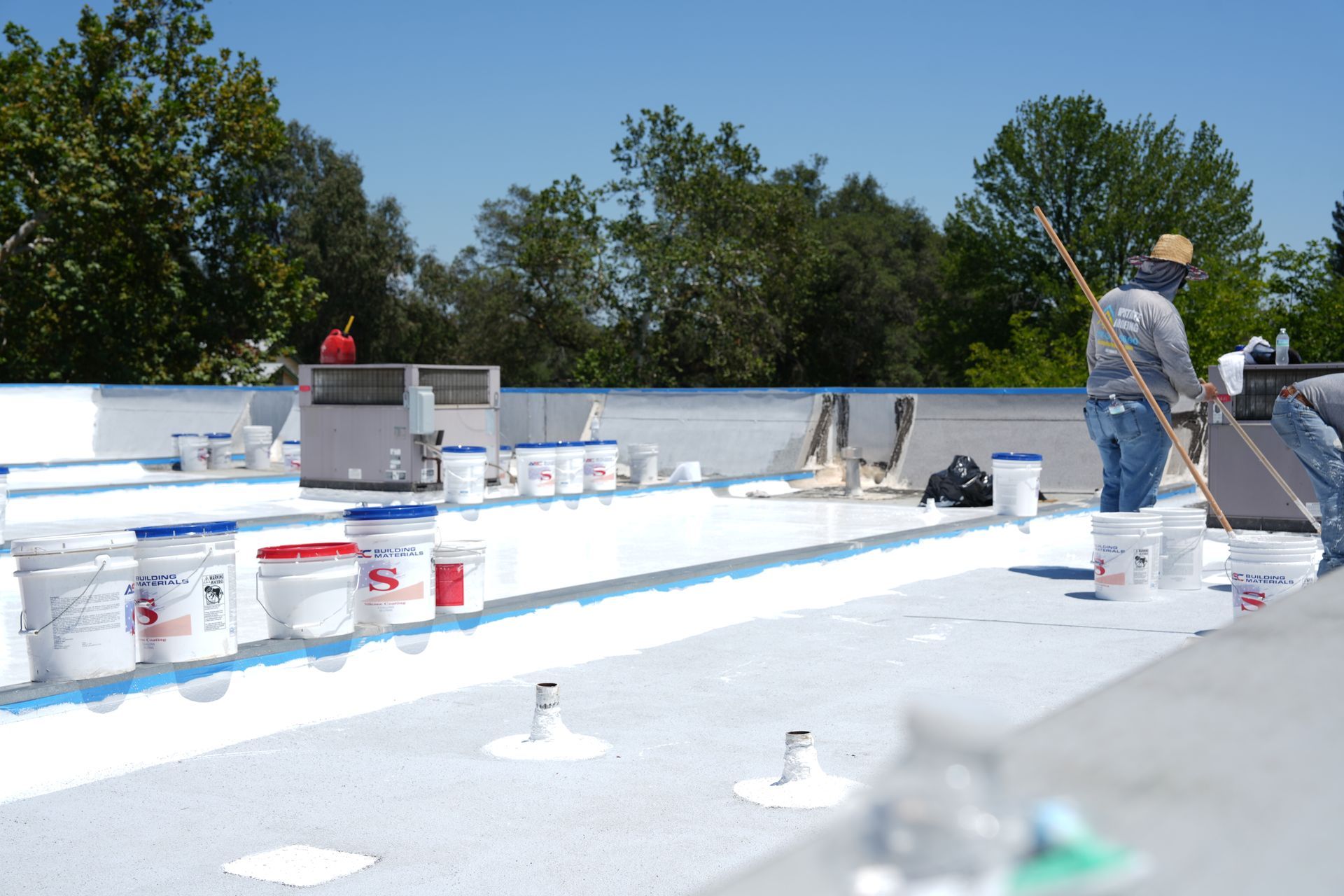 Workers applying white coating on a flat rooftop with paint buckets and trees in the background