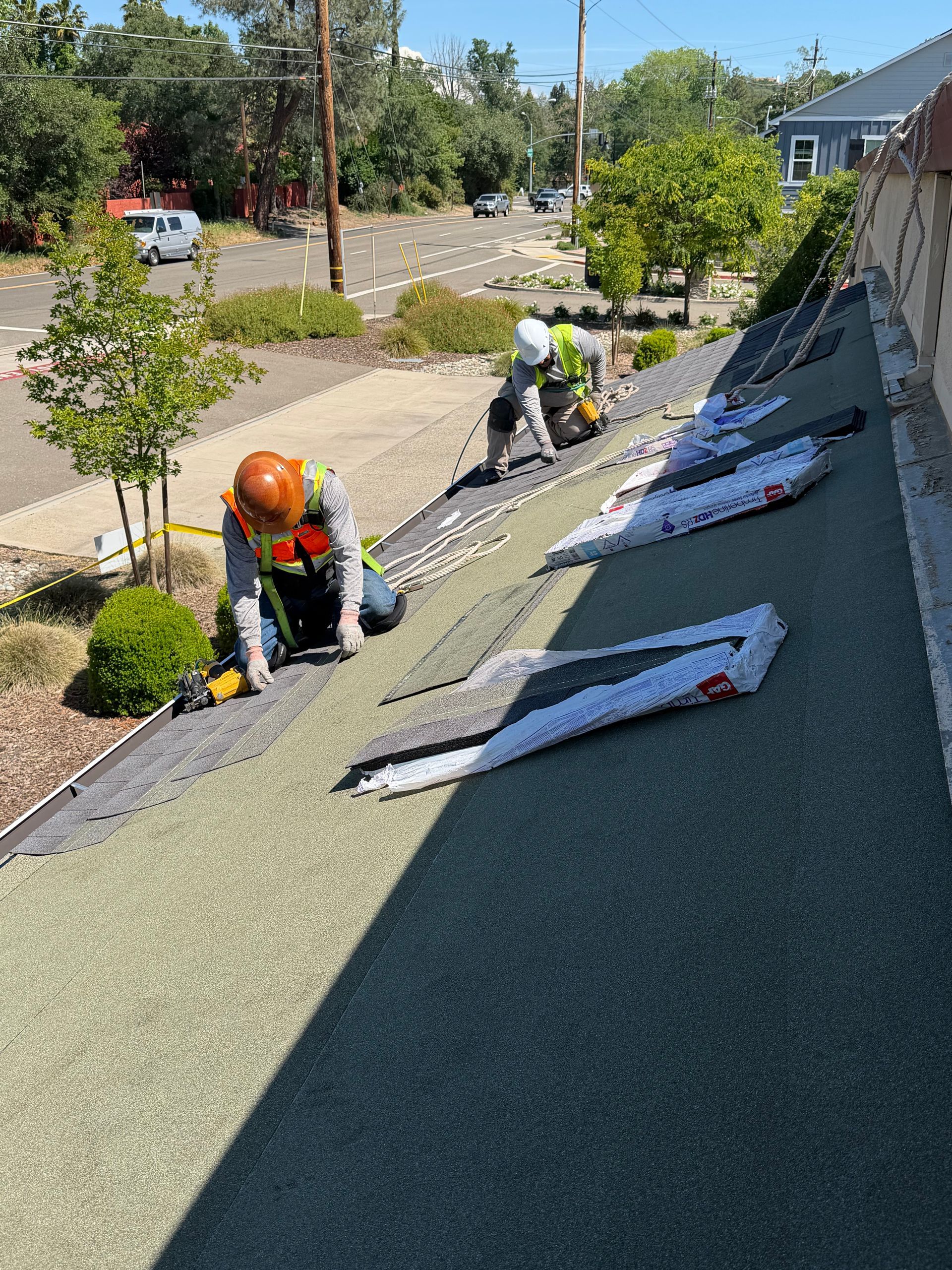 Workers repairing a sloped roof with green shingles, tools and materials laid out on the roof.