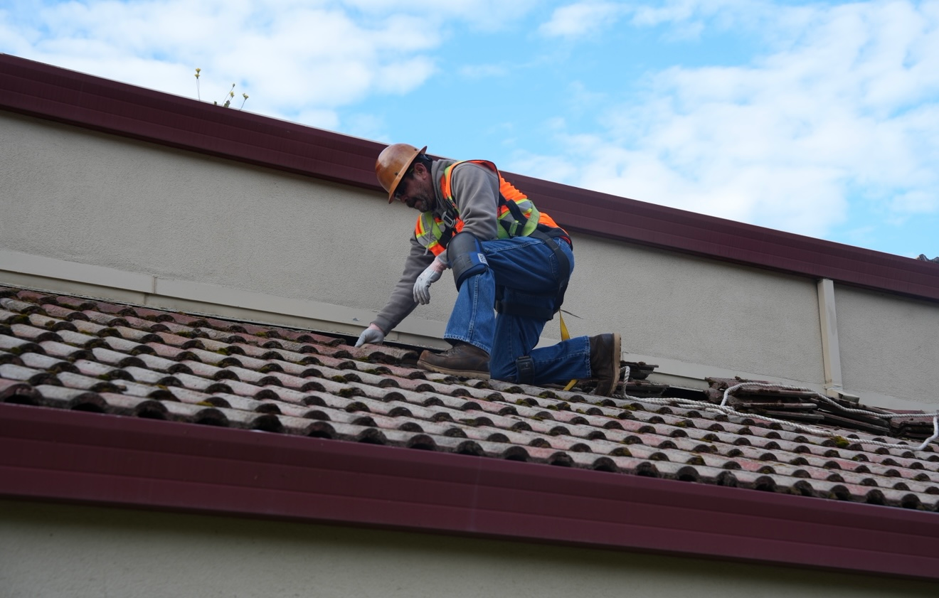 Worker on a steep roof using a safety harness and power tool under a partly cloudy sky