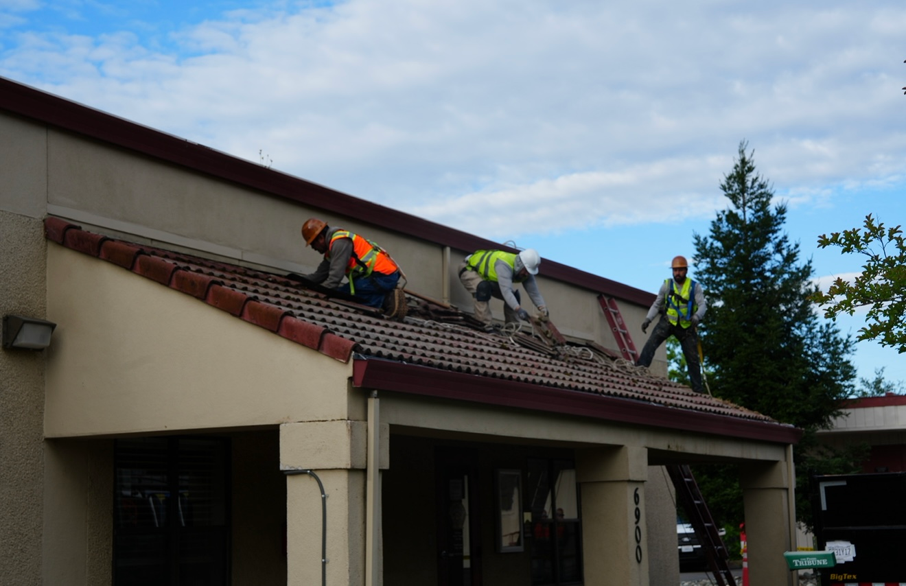 Roofers working on a steep roof of a beige building, wearing safety gear and using ladders.