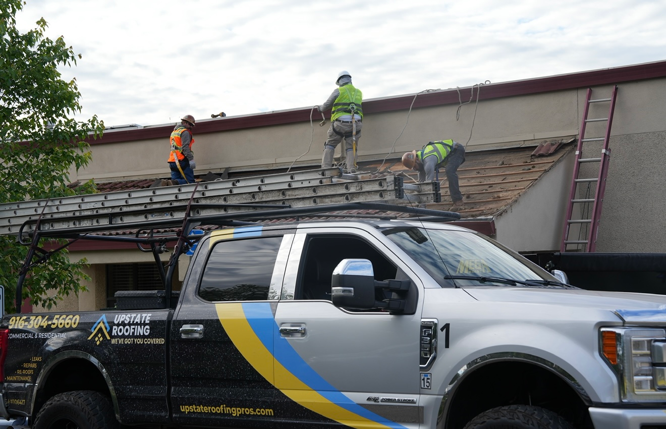 Workers repairing a roof above a white tow truck with a yellow-and-blue stripe.