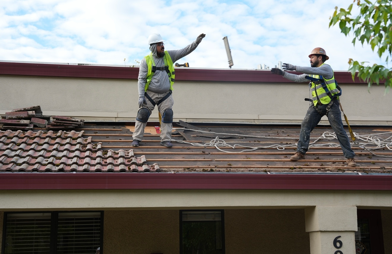 Two roofers in safety vests repair damaged shingles on a house roof.