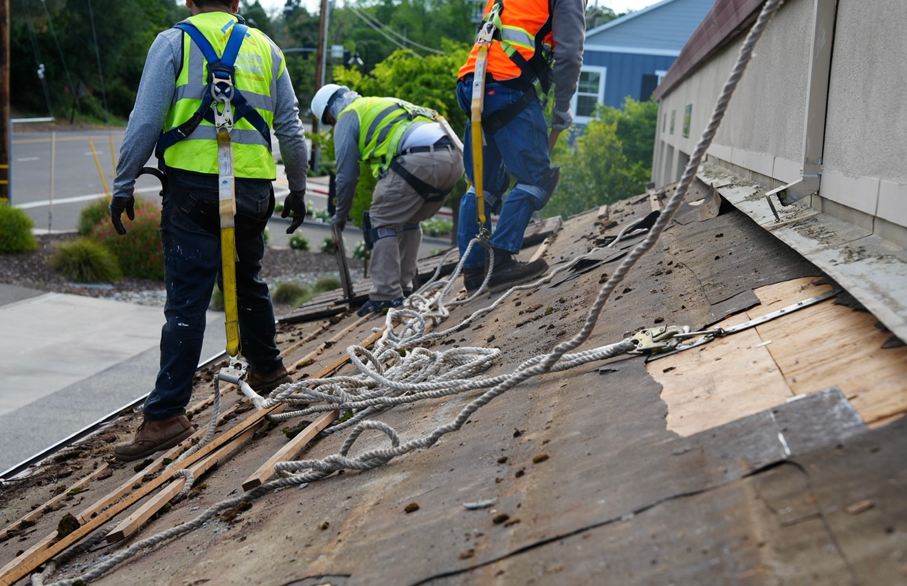 Workers in safety vests repairing a damaged roof with exposed wooden sheathing and debris