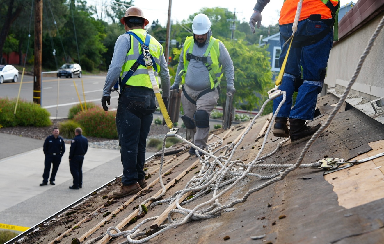 Workers in safety vests repairing a sloped roof with cables and debris nearby