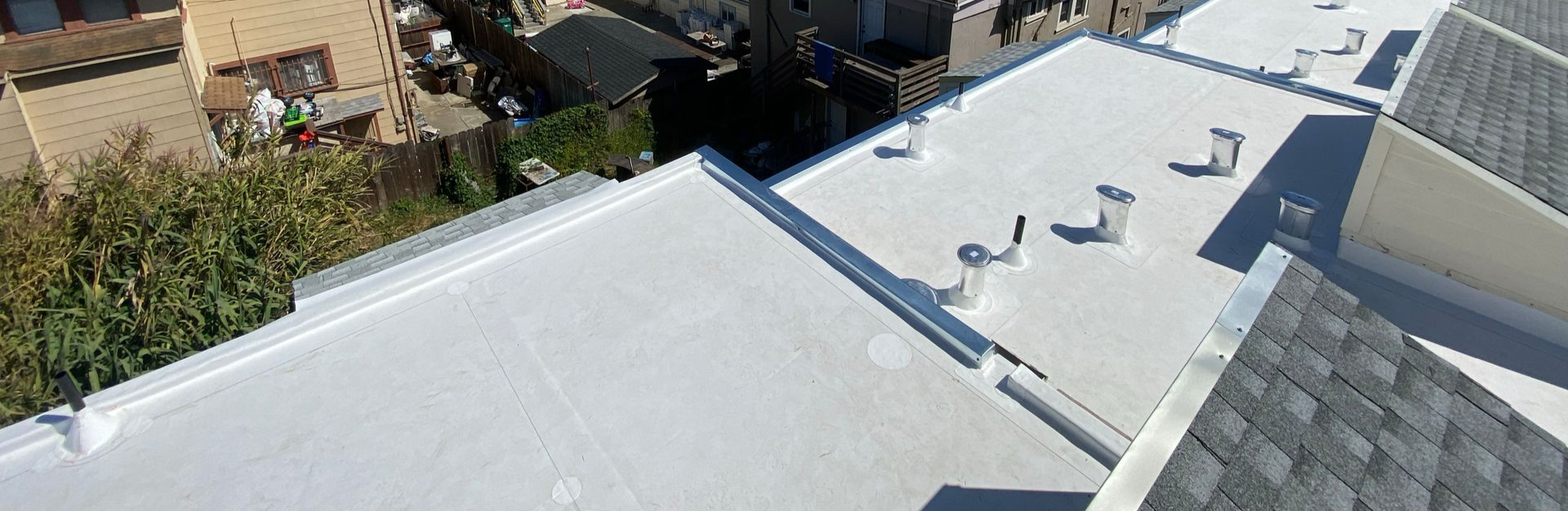 White flat rooftop with skylights and vents beside neighboring buildings and trees