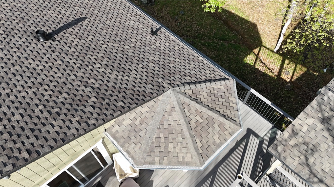 Aerial view of a shingled roof with a small dormer and nearby trees and fence.
