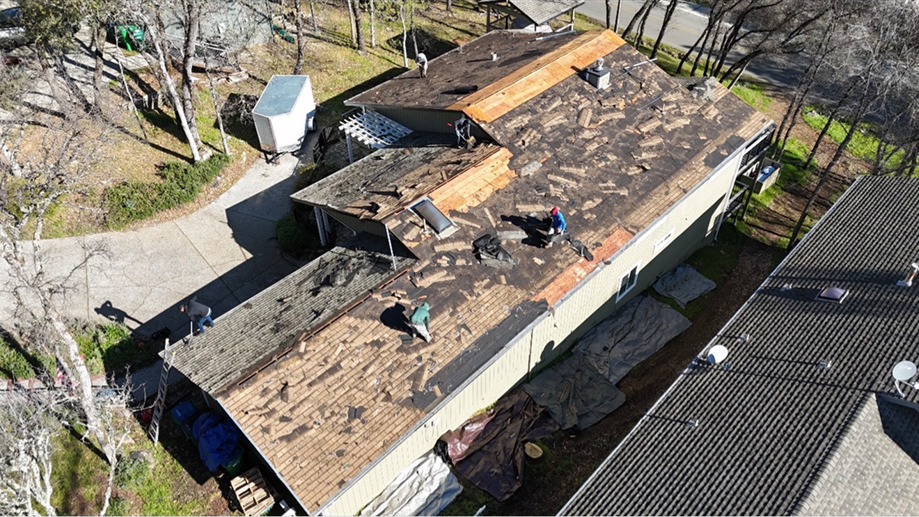 Aerial view of a roof with storm damage and missing shingles, surrounded by trees and neighboring houses